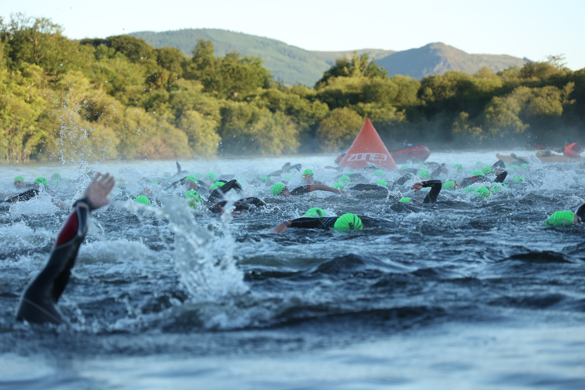 Mass open-water swim start of the Lakesman triathlon, swimmers in green caps with the Lakeland fells behind